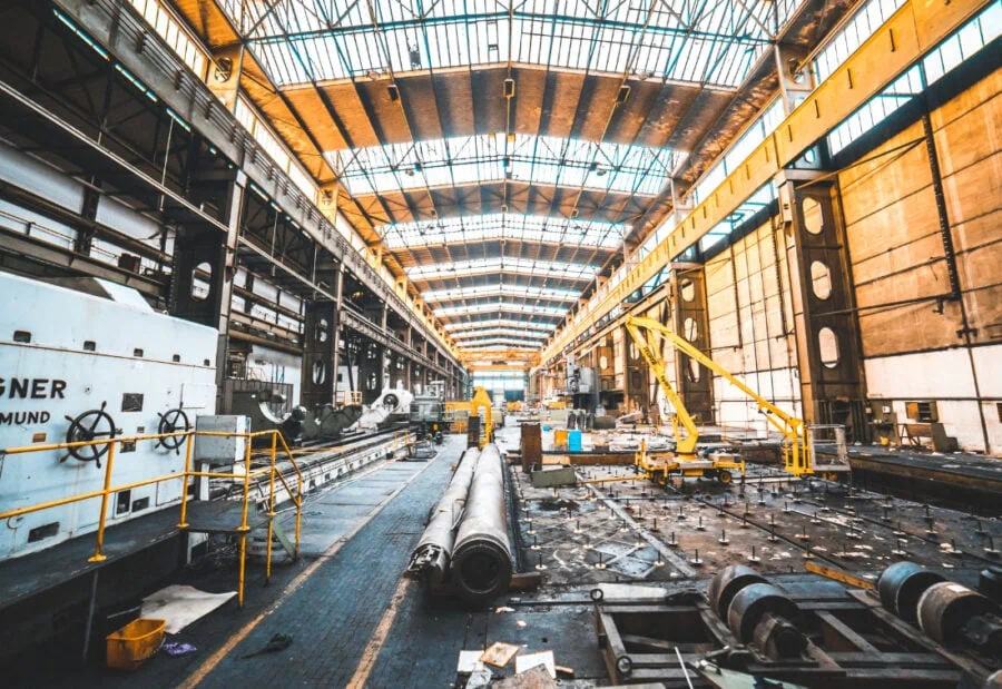 Large, older-style industrial warehouse with heavy equipment, overhead cranes, and long cylindrical metal pieces on the factory floor.